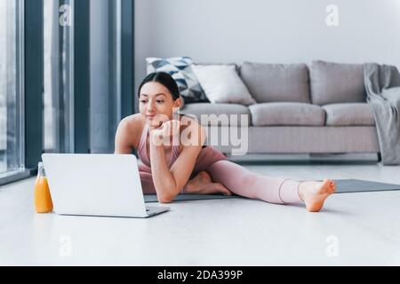 Junge Frau mit schlanker Körperform in Sportswear sitzt auf Matte mit Orangensaft und Laptop drinnen zu Hause Stockfoto