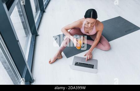 Junge Frau mit schlanker Körperform in Sportswear sitzt auf Matte mit Orangensaft und Laptop drinnen zu Hause Stockfoto