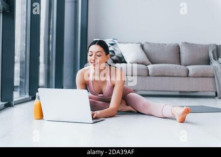 Junge Frau mit schlanker Körperform in Sportswear sitzt auf Matte mit Orangensaft und Laptop drinnen zu Hause Stockfoto