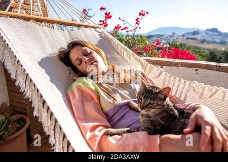 Junge spanische lateinische Frau, die mit ihr in einer weißen Hängematte liegt Tabby Katze auf der Terrasse zu Hause Stockfoto