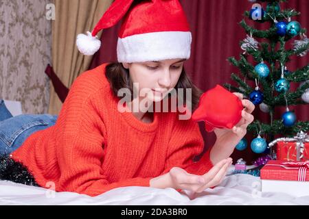 Sparen Sie beim Einkauf von Weihnachtsgeschenken. Trauriges Mädchen in roten weihnachtsmütze und Pullover gießt Münzen aus Sparschwein im häuslichen Interieur Stockfoto