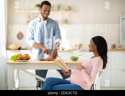 Schwarze schwangere Frau liest Buch, während ihr Mann gesund kocht Frühstück in der Küche Stockfoto