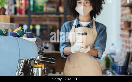 afroamerikanische Hipster Kellnerin in Gesichtsmaske und Handschuhen Hält die Bestellung von Getränken zum Mitnehmen in den Händen, um im Café eine Tasse zu geben Stockfoto