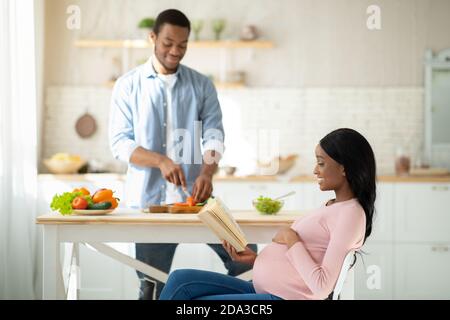 Schwarz erwartungsvolle Dame Buch lesen, während ihr Freund Kochen ausgewogen Frühstück in der Küche Stockfoto