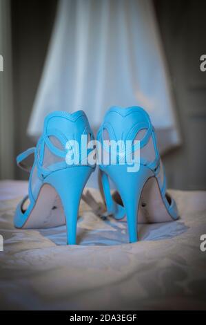 Das weiße Brautkleid der Braut hängt an einem Kleiderschrank mit blauen Schuhen im Vordergrund. Stockfoto