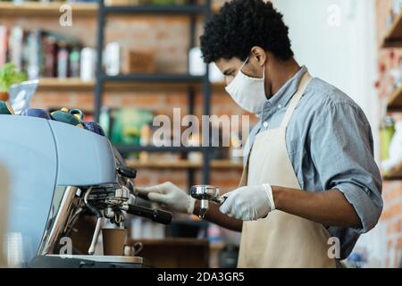 Attraktive Millennial african american Mann in Schürze mit Handschuhen und Schutzmaske macht Espresso Stockfoto