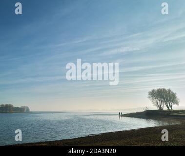 Zwei Männer stehen beim Fischen am Ufer eines Sees, während die Sonne am frühen Morgen an einem Wintertag durch Nebel bricht. Stockfoto