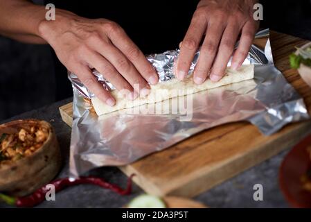 Nahaufnahme eines jungen Mannes, der in Aluminiumfolie ein Durum oder einen Burrito einwickelt, gefüllt mit Hühnerfleisch, das mit verschiedenen Gemüsesorten wie Zwiebeln oder Rot gekocht wird Stockfoto