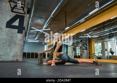 Junge schöne Fitness-Frau in Sportkleidung sitzt auf Yoga-Matte im Fitnessstudio, Blick auf die Kamera, während Sie allein trainieren. Sportliche Menschen, Training, aktive und gesunde Lebensweise Stockfoto