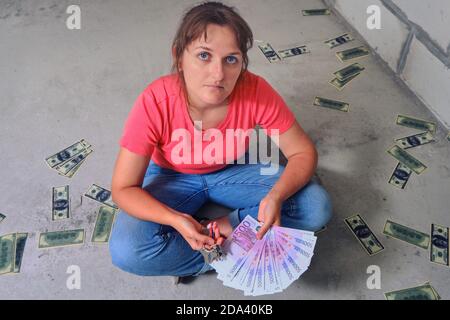 Euro und Dollar unter den Füßen des Mädchens mit den Schlüsseln zur neuen Wohnung. Eine Frau sitzt auf einem leeren Boden mit Geld in den Händen. Stockfoto