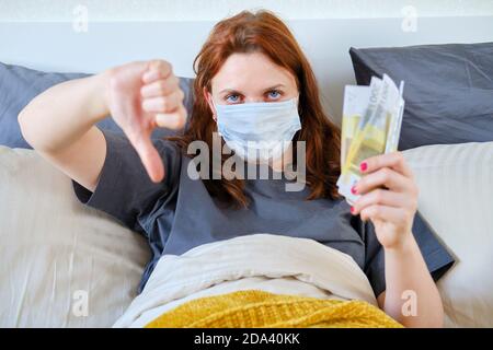 Eine kranke Frau hält Euro-Scheine und zeigt mit dem Finger nach unten eine missbilligende Geste. Ein Mädchen mit einem Coronavirus liegt mit Geld im Bett. Stockfoto