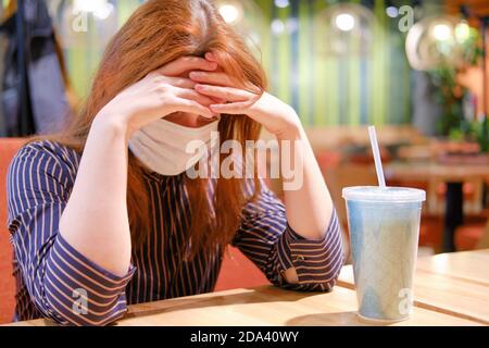 Eine traurige Frau in einer medizinischen Maske sitzt an einem Tisch in einem Café. Probleme mit der Krankheit eines Mädchens, das ihren Kopf in einem Café verneigte. Stockfoto