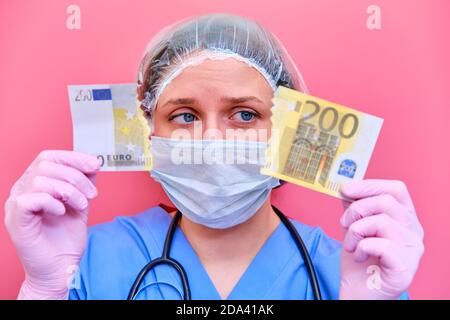 Ein Arzt in blauer Uniform ist unglücklich wegen finanzieller Probleme mit dem Geld. Eine Krankenschwester in einer medizinischen Maske hält eine zerrissene Euro-Banknote auf rotem Hintergrund Stockfoto