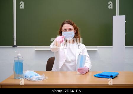 Schularzt in einer medizinischen Maske sitzt an seinem Schreibtisch und zeigt mit dem Finger auf Sie, Kopierraum. Lernschwierigkeiten bei der Grippepandemie Stockfoto