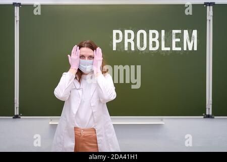 Ein Schularzt in einer medizinischen Maske steht mit einem traurigen Gesicht. Textproblem ist in Kreide auf der Tafel geschrieben Stockfoto