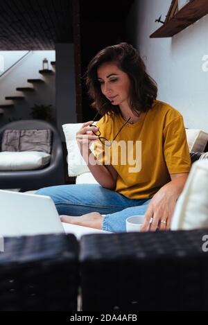 Brunette Frau arbeitet von zu Hause mit Laptop hält eine Tasse Kaffee. Stockfoto