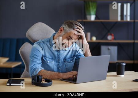 Müde und enttäuscht Senior Mann arbeitet im Home Office mit Laptop. Porträt eines hübschen grauhaarigen Managers, der am Arbeitsplatz sitzt. Online studieren Stockfoto