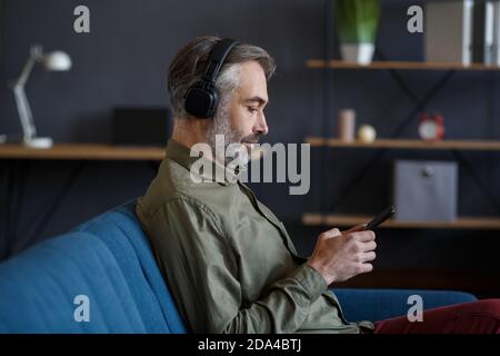 Älterer Mann mit Kopfhörern, der Musik auf dem Smartphone über die Musik-App hört. Porträt eines schönen grauhaarigen Mannes in Kopfhörer und Handy zu Hause Stockfoto