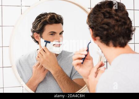 Junger Mann, der auf den Spiegel schaut, während er sich auf verschwommenem Vordergrund rasiert Im Badezimmer Stockfoto