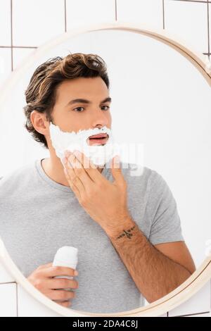 Junger Mann, der Rasierschaum in der Nähe des Spiegels im Badezimmer anwendet Stockfoto