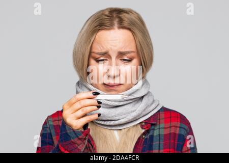Ungesunde Frau erkältet und Blick auf Thermometer. Verärgert Frau im Schal, kariertes Hemd fühlt sich schlecht. Kälte, Krankheit. Stockfoto