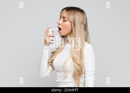 Verärgert junge blonde Frau mit Spray, um Halsschmerzen zu behandeln. Rachenschmerzen, schmerzhaftes Schluckkonzept Stockfoto