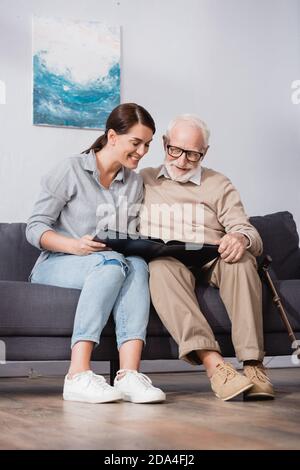 Fröhlicher Vater und Tochter beim Stöbern im Fotoalbum, während sie darauf sitzen Sofa zu Hause Stockfoto