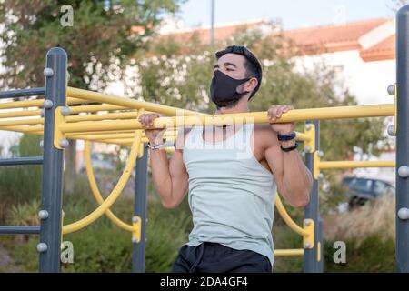 Junger Latino-Mann in hellblauem Tank Top und schwarz Hose und Maske machen Übungen in einem Calisthenics Park Stockfoto