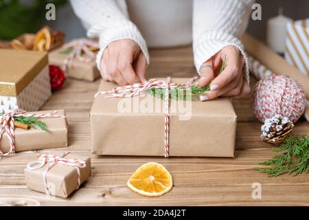 Frau s Hände Verpackung Weihnachtsgeschenk, Nahaufnahme. Unvorbereitete Geschenke auf Holzhintergrund mit Dekorelementen und Gegenständen, Draufsicht. Weihnachten oder Neues Ye Stockfoto