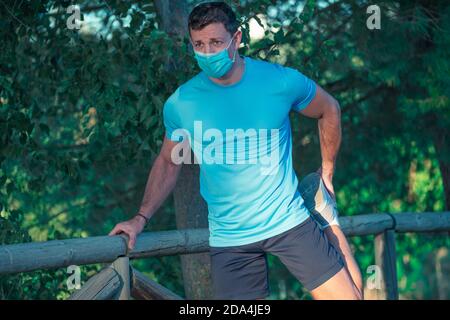 Junger Kaukasianer mit Maske und blauem Hemd beim Dehnen Auf dem Zaun eines Parks mit Bäumen in der Hintergrund Stockfoto