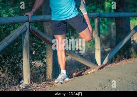 Junger Kaukasianer mit Maske und blauem Hemd beim Dehnen Auf dem Zaun eines Parks mit Bäumen in der Hintergrund Stockfoto