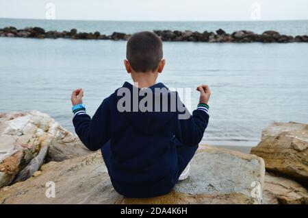 Junger Mann meditiert während des Sonnenuntergangs auf der Klippe des Ozeans. Der Junge macht Yoga in einem dunkelblauen Trainingsanzug. Sitzt auf den Felsen am Meer. Stockfoto