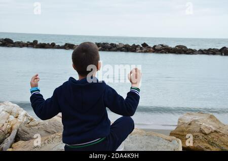 Junger Mann meditiert während des Sonnenuntergangs auf der Klippe des Ozeans. Der Junge macht Yoga in einem dunkelblauen Trainingsanzug. Sitzt auf den Felsen am Meer. Stockfoto