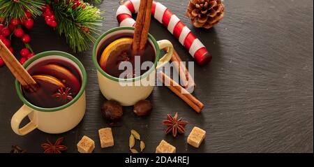 Glühwein, winterwärmendes Alkoholgetränk, zwei Jahrgangsbecher, Zutaten Orange und Gewürze und Weihnachtsdekoration auf schwarzem Stein, Nahaufnahme. Stockfoto