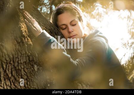 Close Portrait der jungen Frau umarmt den Stamm eines Alte Eiche mit Sonneneinstrahlung im Hintergrund Stockfoto