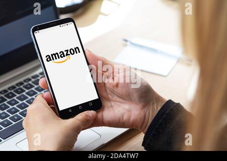 mazon-Logo auf dem Bildschirm des Mobiltelefons. Blonde Frau an ihrem Arbeitsplatz, vor dem Laptop mit Smartphone in Stockfoto