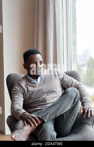 Selbstbewusster, stilvoller schwarzer Mann, der zu Hause im Stuhl sitzt und durch das Fenster schaut. Stockfoto
