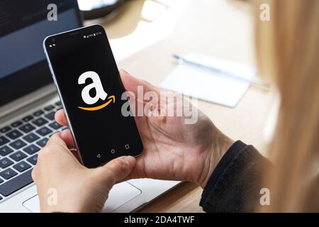 mazon-Logo auf dem Bildschirm des Mobiltelefons. Blonde Frau an ihrem Arbeitsplatz, vor dem Laptop mit Smartphone in Stockfoto