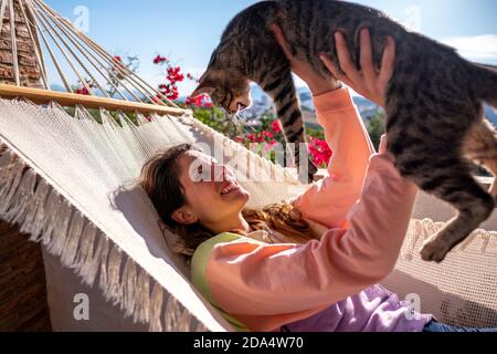 Junge spanische lateinische Frau, die mit ihr in einer weißen Hängematte liegt Tabby Katze auf der Terrasse zu Hause Stockfoto