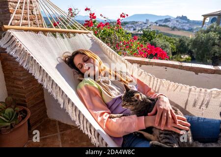 Junge spanische lateinische Frau, die mit ihr in einer weißen Hängematte liegt Tabby Katze auf der Terrasse zu Hause Stockfoto