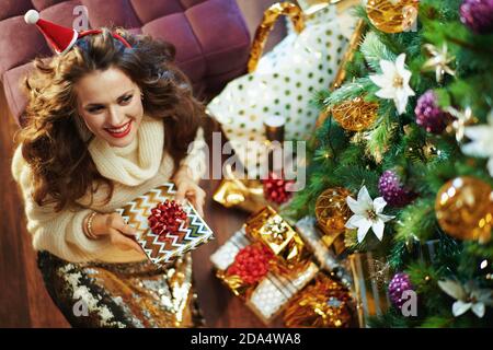 Frohe Weihnachten. Obere Ansicht der glücklich trendy mittleren Alters Hausfrau in Gold Pailletten Rock und Pullover mit Weihnachtsgeschenk Box in der Nähe Weihnachtsbaum und Stockfoto