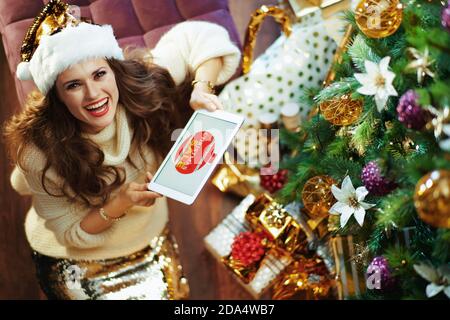 Frohe Weihnachten. Obere Ansicht der glücklich stilvolle Frau mittleren Alters in Gold Pailletten Rock und Pullover mit Weihnachtscoupon auf einem Tablet-PC in der Nähe von Weihnachten tr Stockfoto