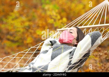 Entspannte Frau, die Schutzmaske trägt, die auf Hängematte in einem ruht Wald in der Herbstsaison Stockfoto