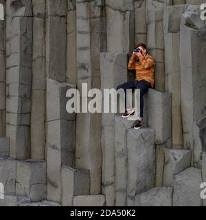 Mann, der auf Basaltfelsen sitzt und mit der Kamera fotografiert, Reynisfjara Black Sand Beach, Vik, Myrdalshreppur, Southern Region, Island Stockfoto