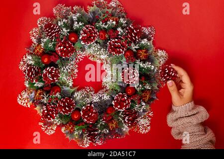 Eine Frauenhand in einem beigen Pullover macht einen Weihnachtskranz aus Zapfen, Beeren und Kugeln, isoliert auf einem roten Hintergrund Stockfoto