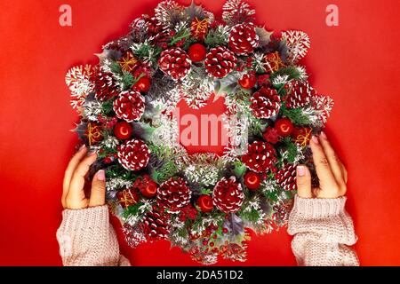 Zwei Frauenhände in einem Strickpullover halten einen Weihnachtskranz aus Zapfen, Beeren und Kugeln auf einem leuchtend roten Hintergrund Stockfoto