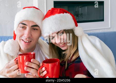 Weihnachtsstimmung. Junge glückliche Paar trinken heißen Tee oder Kakao sitzen zu Hause auf dem Sofa tragen weihnachtsmützen Stockfoto