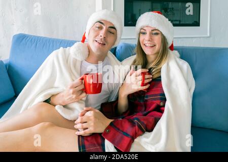 Junge kaukasische Paar trinken heißen Tee oder Kakao aus rot Tassen, während Sie zu Hause auf dem Sofa sitzen und den weihnachtsmann tragen Hüte und Schlafanzug in einem weißen bla gewickelt Stockfoto