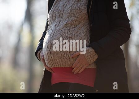 Nahaufnahme einer schwangeren Frau, die ihren geschwollenen Bauch in Winterkleidung berührt, die draußen in der Natur steht. Stockfoto