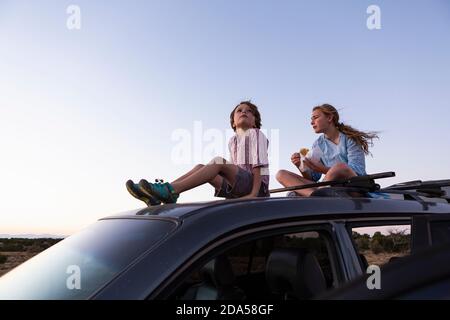 Teenager-Mädchen und ihr jüngerer Bruder sitzen auf ihrem SUV bei Sonnenuntergang. Stockfoto